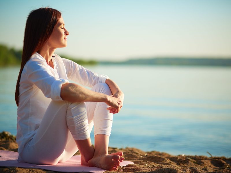 Side view of serene woman sitting on sandy beach against blue sky outdoors